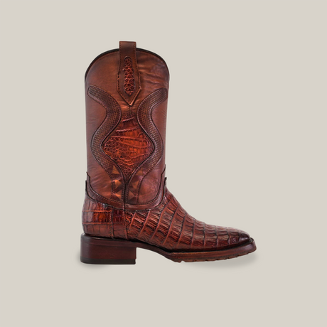 The Exotic Caiman Belly Tail boot in brandy features a textured crocodile leather design, high shaft with decorative stitching, and a low stacked heel. Displayed against a white background, it embodies the classic charm of Western fashion with a double sole square toe.