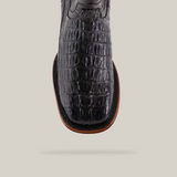 Close-up of the Exotic Caiman Tail - Diamond - Black cowboy boot with a square toe, featuring a black crocodile pattern. Its polished finish and brown sole add elegance against a plain light beige background.