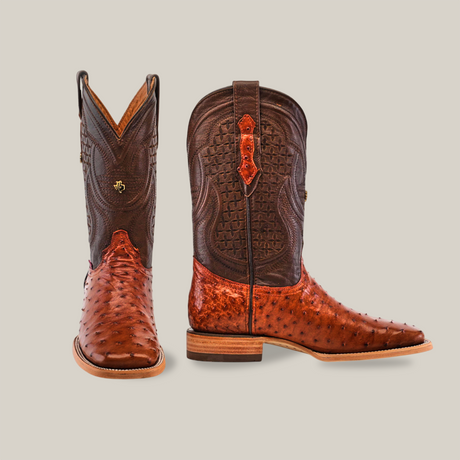 The Exotic Full Quilt Ostrich - Brandy - Square Toe boots embody the cowboy lifestyle with their handcrafted elegance, offering rich brown tones, intricate stitching, and a classic Western design—one boot stands tall while the other lays on its side.