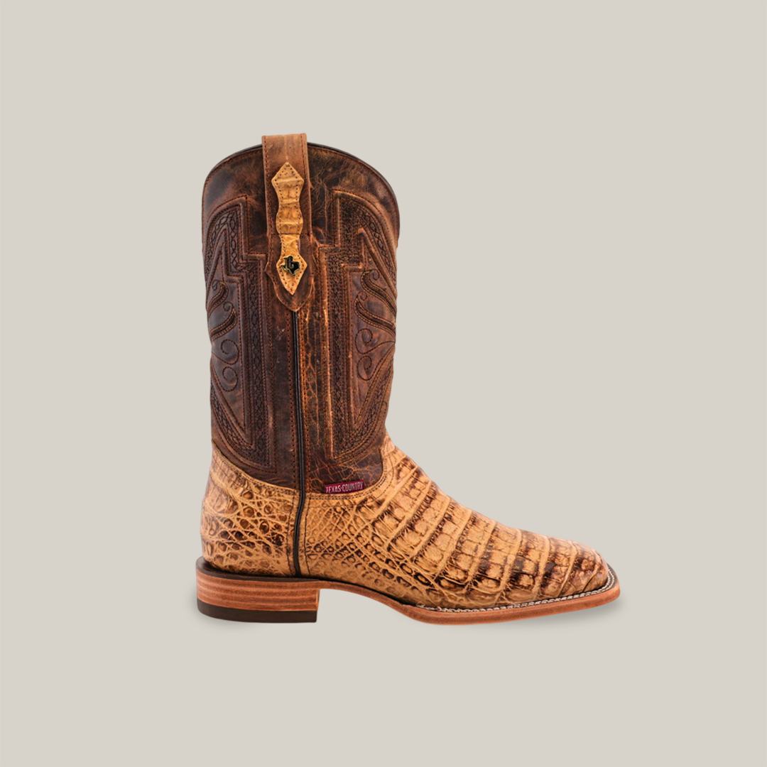 The Exotic Caiman Belly - Antique - Square Toe cowboy boot features tan and brown hues with intricate patterns, complemented by a wooden sole and small side logo. Its authentic western look comes from the unique textures and colors of the Exotic Caiman Belly design.