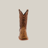 Front view of a single, square-toed brown cowboy boot named Exotic Caiman Horn Back - Antique, showcasing intricate shaft stitching and a textured caiman-like pattern on the foot, with a handcrafted slightly curved top and low heel against a plain backdrop.