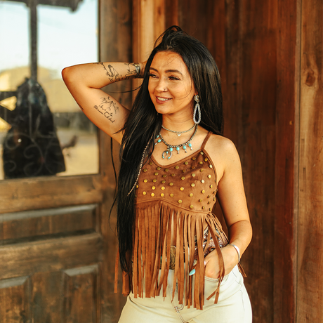 A woman poses before a wooden wall, smiling and holding her long black hair. She wears the LB Brown Studded Suede Crop T0156-BRW with white pants and turquoise jewelry, exuding boho-chic elegance. Her left arm features tattoos, complementing the Western-style rustic door backdrop.