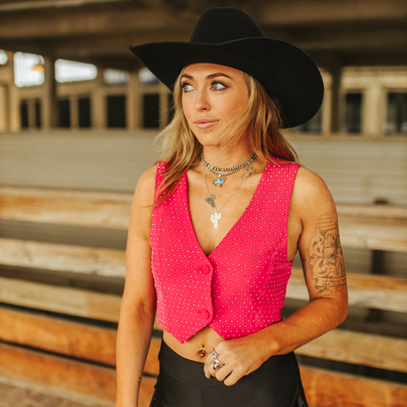 A woman in a LB Hot Pink Bling Vest and black cowboy hat stands outside, highlighting her feminine silhouette. Her long hair drapes over tattooed arms, paired with layered necklaces and rings. Rhinestone embellishments sparkle against the wooden structure behind her.