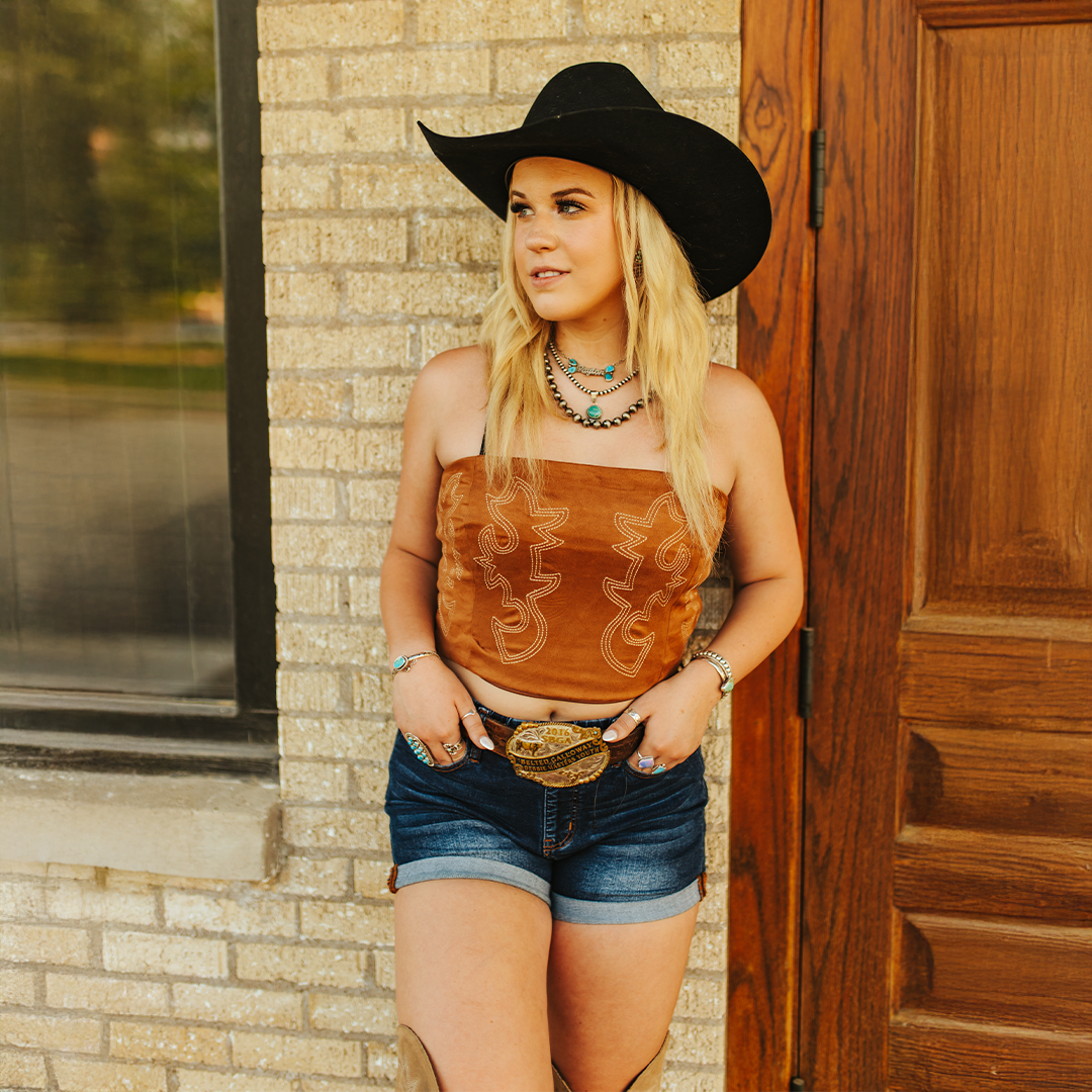 A woman in an LB Camel Corset Cream Embroidery T0655-CML and denim shorts leans against a brick wall wearing a black cowboy hat and boots. She accessorizes with turquoise jewelry and a large belt buckle, gazing to the side with a wooden door in the background.