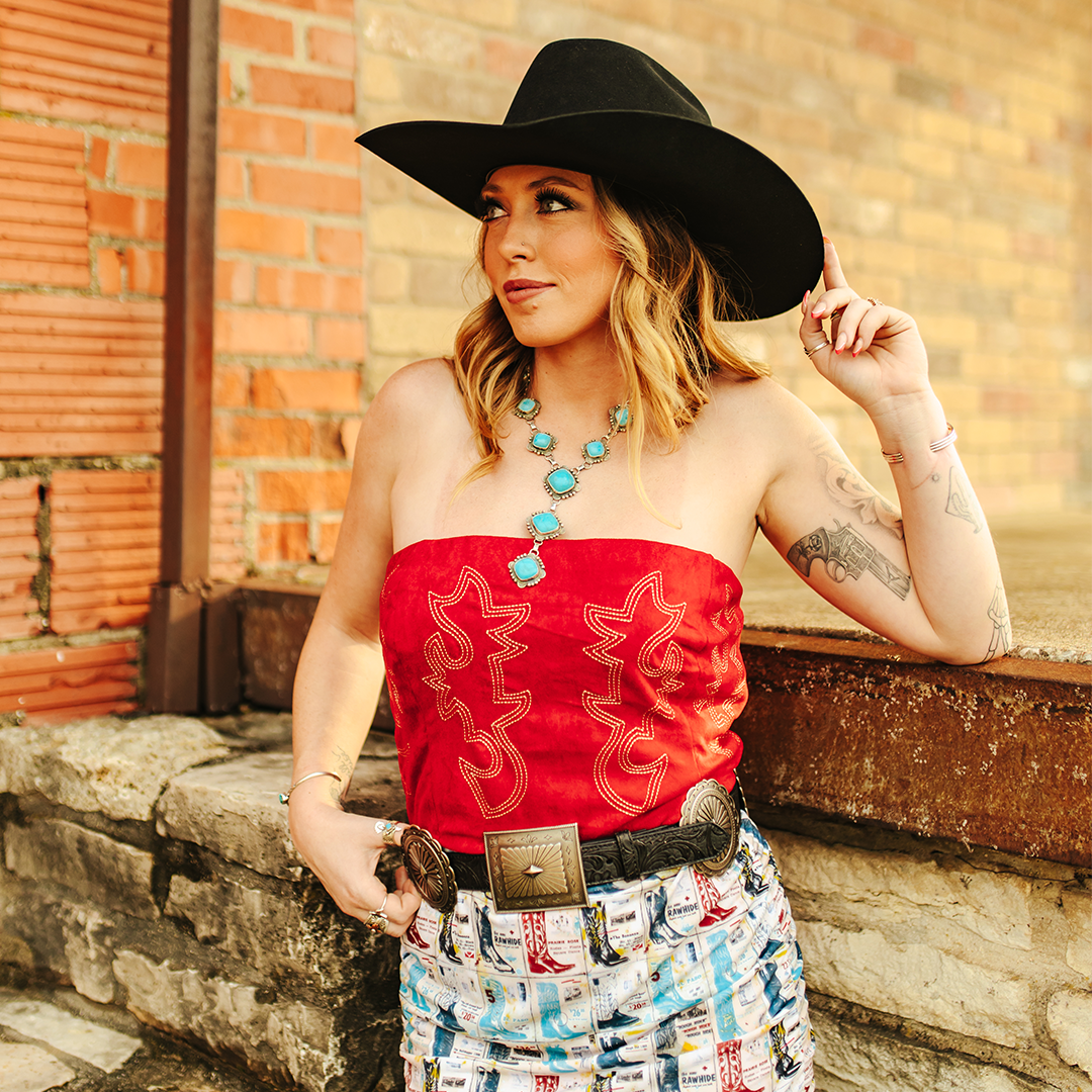 A woman in a black cowboy hat and the LB Red Corset Cream Embroidery T0655-RED with adjustable back lacing stands against a brick wall. Shes adorned with turquoise jewelry and has tattoos on her arms, set against a rustic charm of weathered bricks and stone.