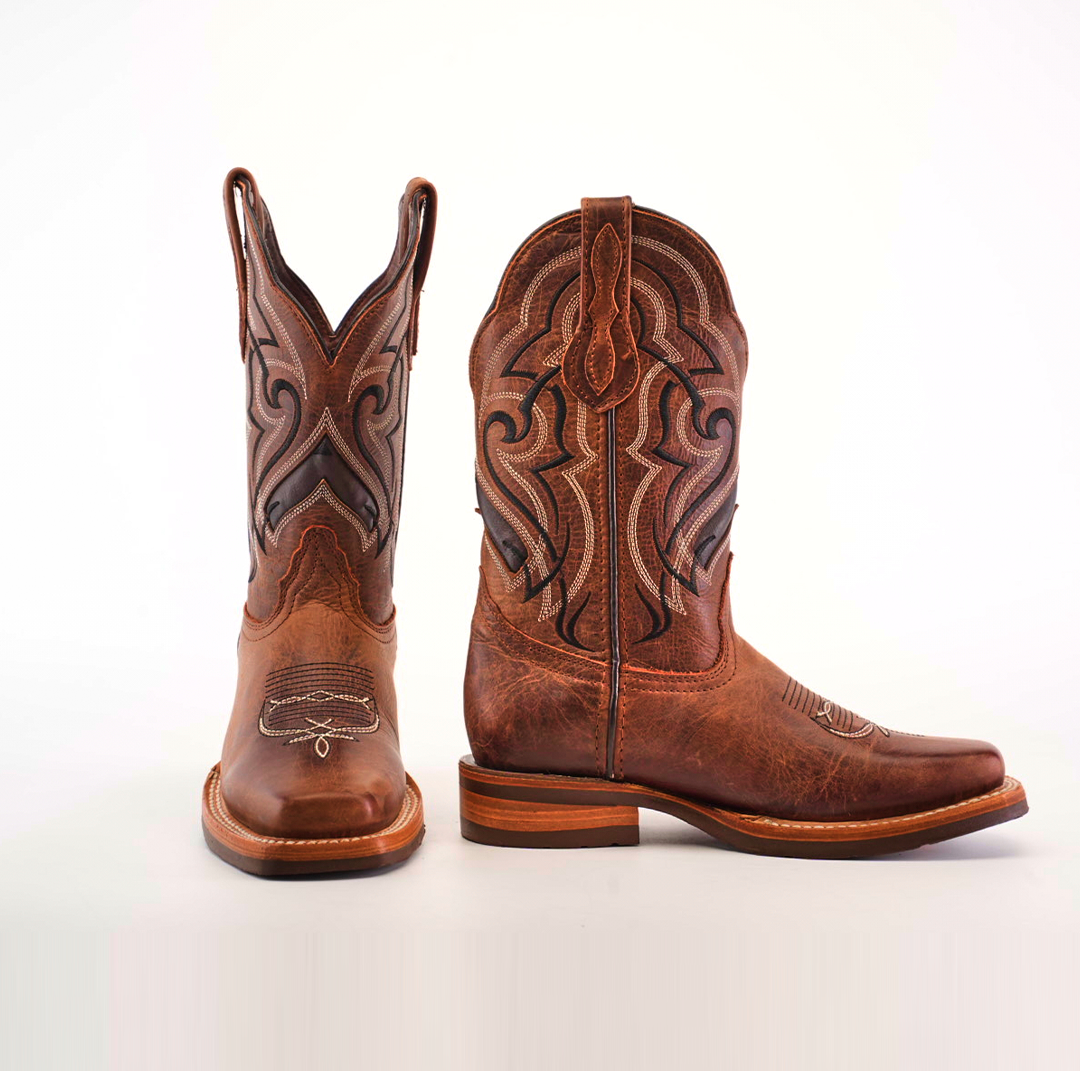 The Fresno Shedron boots feature a stylish rodeo toe with intricate black and brown stitching on the shafts. One boot stands upright while the other is angled to reveal its side pattern and slip-resistant sole, all set against a white background.