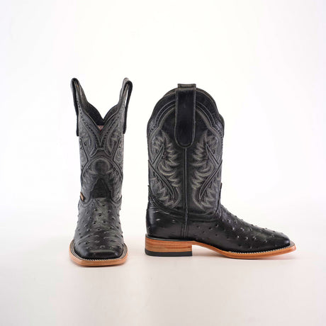 The Karoo Ostrich Print Black Square Toe boots feature a luxurious design with intricate patterns, a smooth, shiny texture, and exotic ostrich print. Displayed against a white backdrop, one boot stands upright while the other lies on its side, showcasing decorative stitching and a light brown sole.