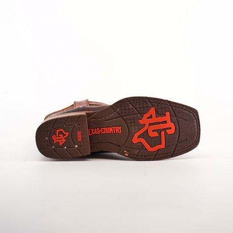 The image shows the sole of an Azkar Moka Slip Resistant Sole Rodeo Toe boot, adorned with an orange longhorn logo and TEXAS COUNTRY in bright text. The detailed tread provides slip resistance, and the brown cowboy boot rests on a clean white surface.