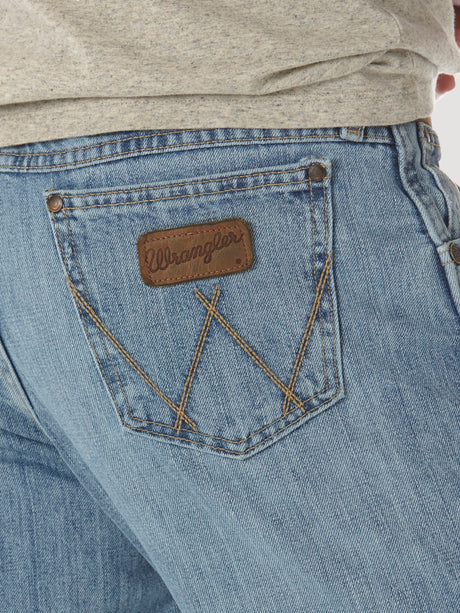 A detailed image shows the back pocket of Mens Wrangler Retro Relaxed Fit Bootcut Jean - WRT20CR in light blue, featuring brown stitching and a leather patch with the brand name. Above the jeans, there is a partially visible gray shirt.