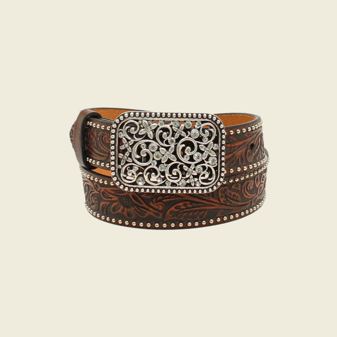 The Ariat Western Girls Belt (Youth Leather, A1303602) features ornate brown leather with intricate floral and studded designs, embodying Western inspiration. Its silver buckle is embellished with swirls and gemstone-like accents. The background is plain off-white.