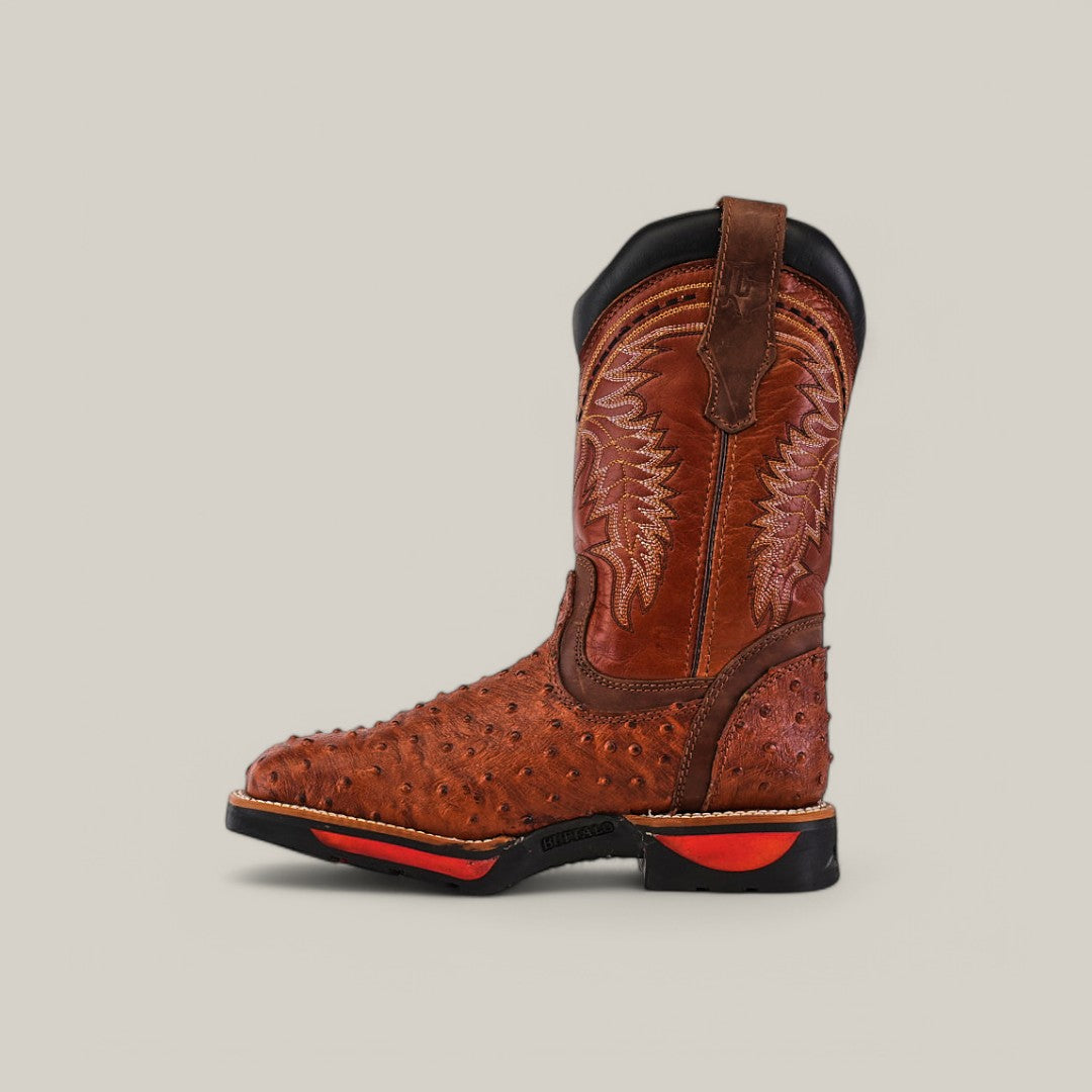 The Ostrich Print Cogñac Composite Toe - E6022 is a single brown cowboy boot with a textured ostrich print and intricate shaft stitches. Designed by Texas Country Work Boots, it features a black and red sole, pull strap, and durable style against a light grey background.