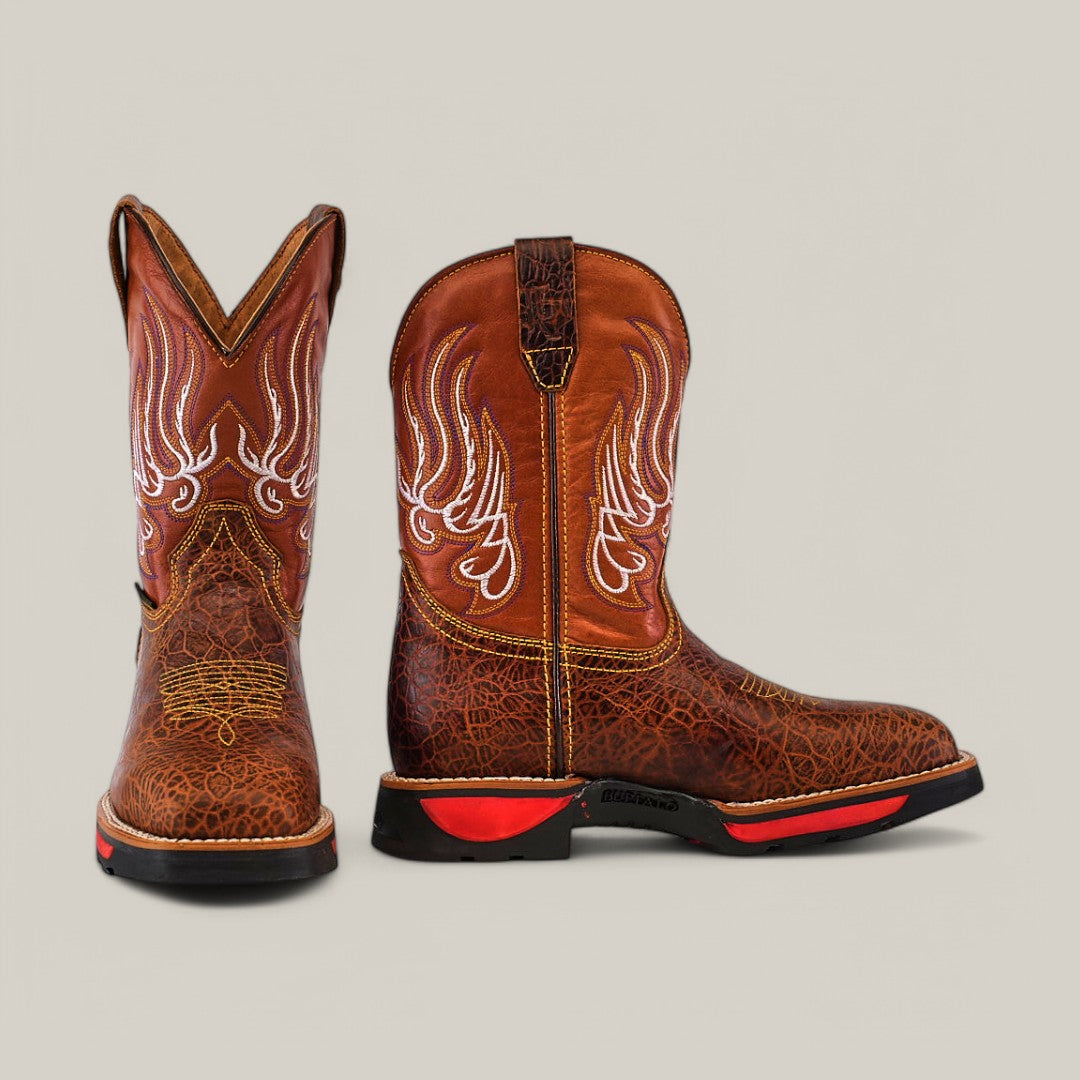 A pair of Jurasic Honey Soft Toe - E6080 boots, with intricate white stitching, is set against a neutral backdrop. The durable brown cowboy boots boast black and red soles, offering a rugged and textured appearance.