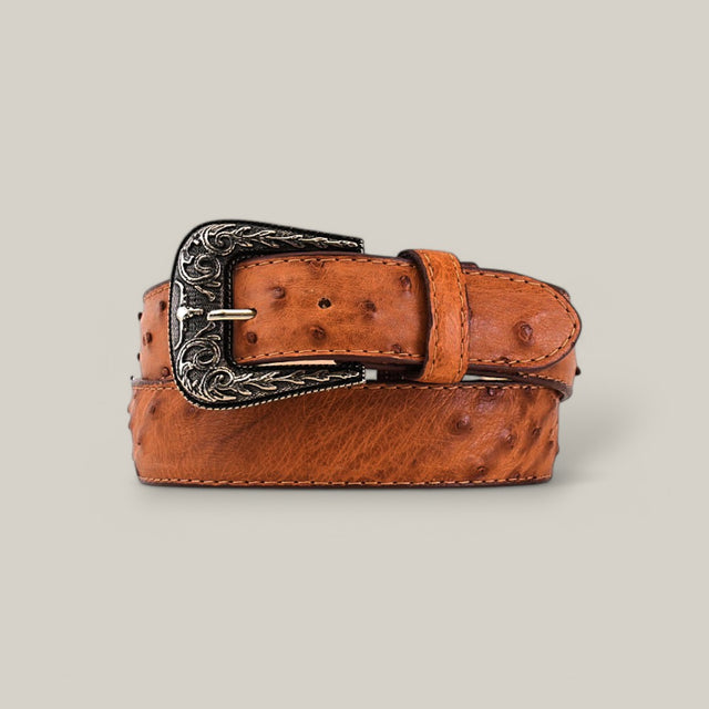 A rolled-up Exotic Full Quilt Ostrich Brandy Belt features premium ostrich leather and a textured surface, highlighted by a Texas Buckle with intricate silver engravings, against a plain beige background.