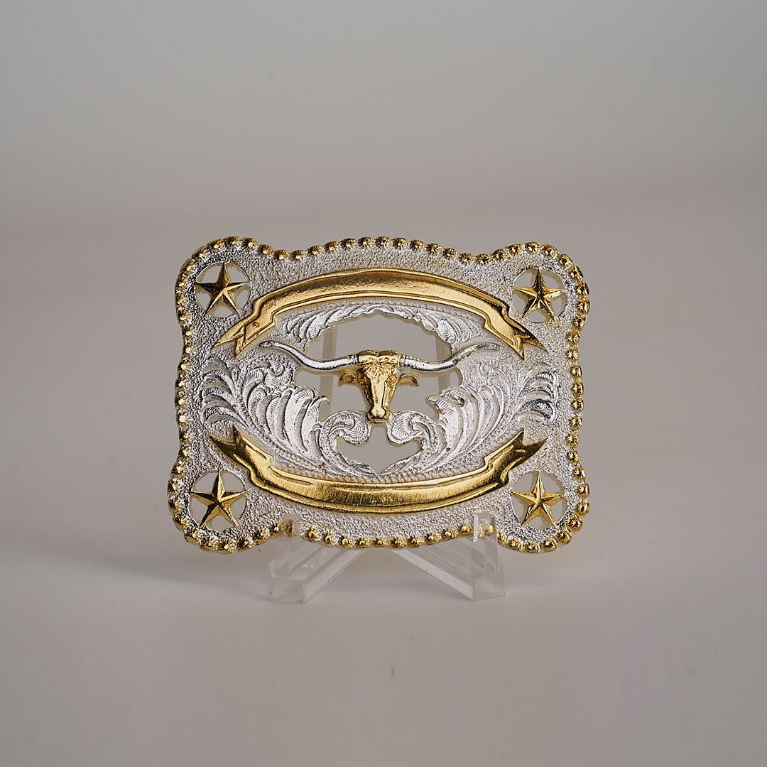 The LongHorn Buckle is a rectangular, Texas-style accessory made from alpaca silver, featuring ornate gold detailing with a central bulls head. Gold stars adorn the corners within a beaded border, all set against a neutral background that highlights its western charm.