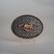 The Racing Horse Buckle is an oval belt accessory featuring a gold horse design at its center amidst intricate geometric patterns, reminiscent of classic Texas buckles. It includes small gold accents along the edge and is displayed on a clear stand against a neutral background.