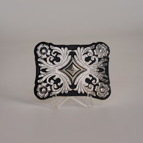 The Floral Diamond Buckle, an ornate Texas-style piece crafted from alpaca silver, features intricate floral and scroll designs. It is elegantly displayed on a small transparent stand against a plain background.