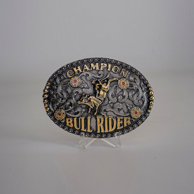 The Bull Riding Champion Buckle showcases timeless western designs with an ornate oval shape and Champion Bull Rider text. It features an embossed bull rider image, intricate detailing, and decorative red gemstone accents on an alpaca silver background, ideal for Texas-style enthusiasts.