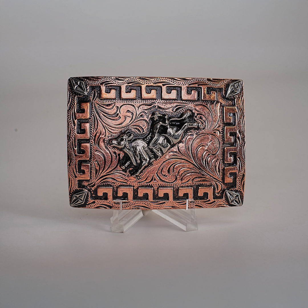 The Bull Rider Buckle is a decorative rectangular belt buckle with western designs, swirling motifs, geometric border patterns, and an engraved bull at the center. It features a copperish tone with darker accents and is showcased on a clear stand.