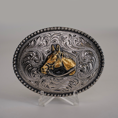 The Stud Horse Buckle is an ornate, oval-shaped Texas-style creation in alpaca silver, featuring intricate western scrollwork with a central gold horse head design against a neutral background.