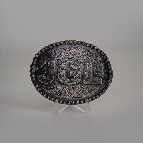 The JGL Buckle, an intricately designed oval piece made from alpaca silver, showcases bold JGL letters and ornate swirling patterns like Texas-style buckles. It is displayed on a clear stand against a plain backdrop.