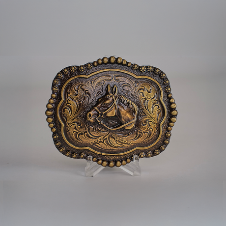 The Vintage Stud Horse Buckle in alpaca silver displays an embossed horse head and floral designs on a small stand. A beaded border enhances its shimmer in bronze and gold against a gray background.