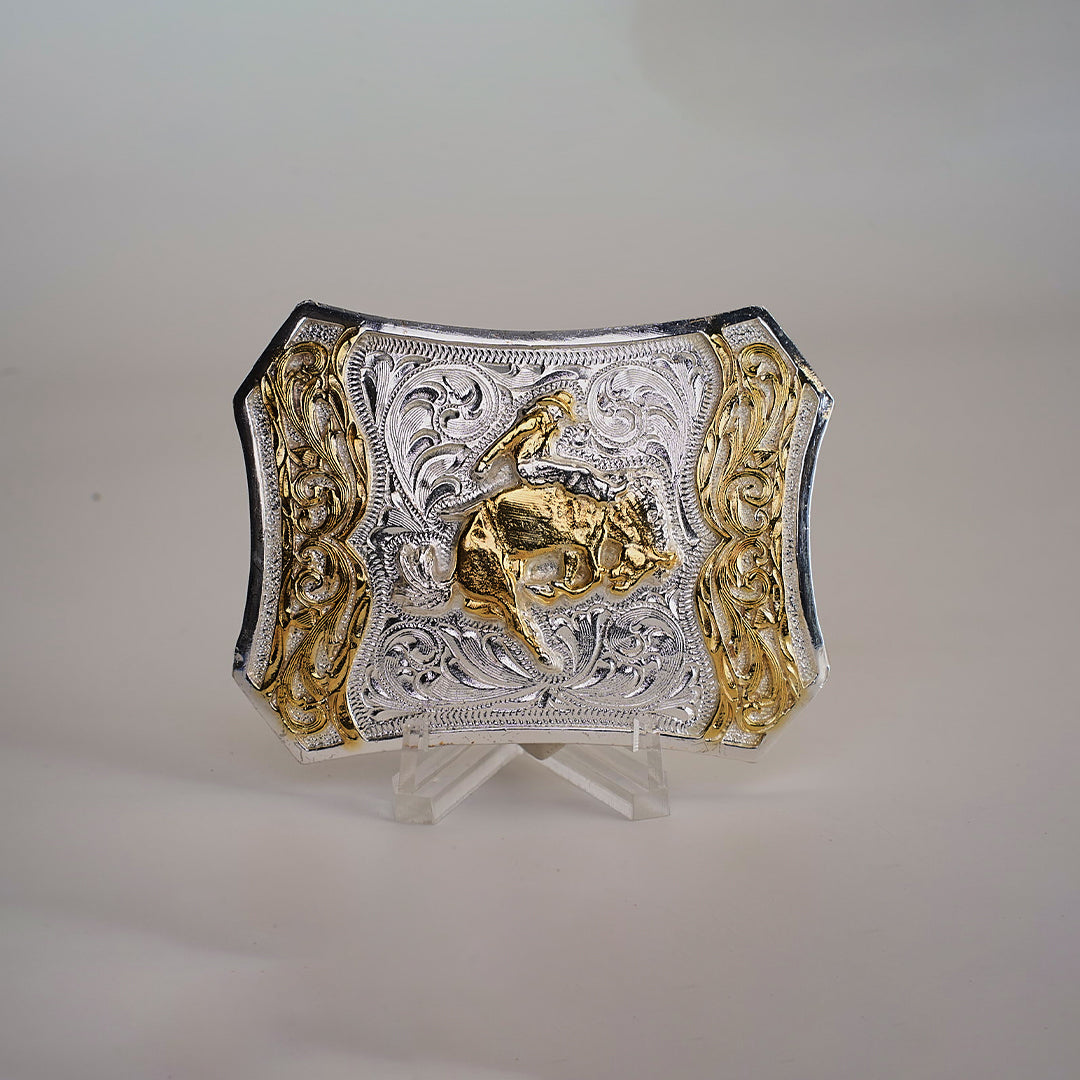 The Spade Bull Rider Buckle is a Texas-style piece crafted from alpaca silver. It features intricate engravings of a cowboy riding a bull, with silver and gold accents that pop against its plain background, showcasing the detailed western design.