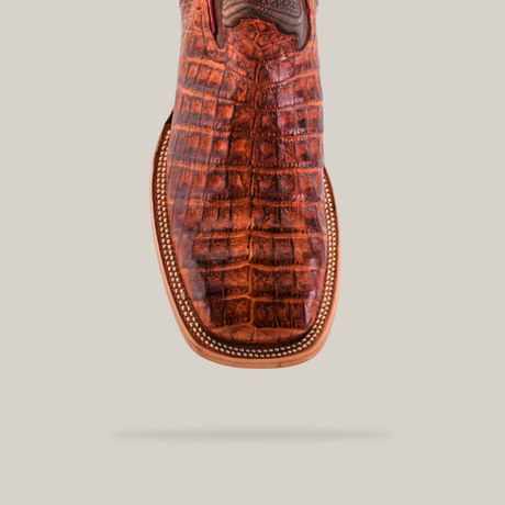 A close-up of the square toe of an Exotic Caiman Belly boot in Brandy, crafted from reddish-brown caiman leather with a crocodile pattern. The boot features decorative stitching, set against a light beige background.