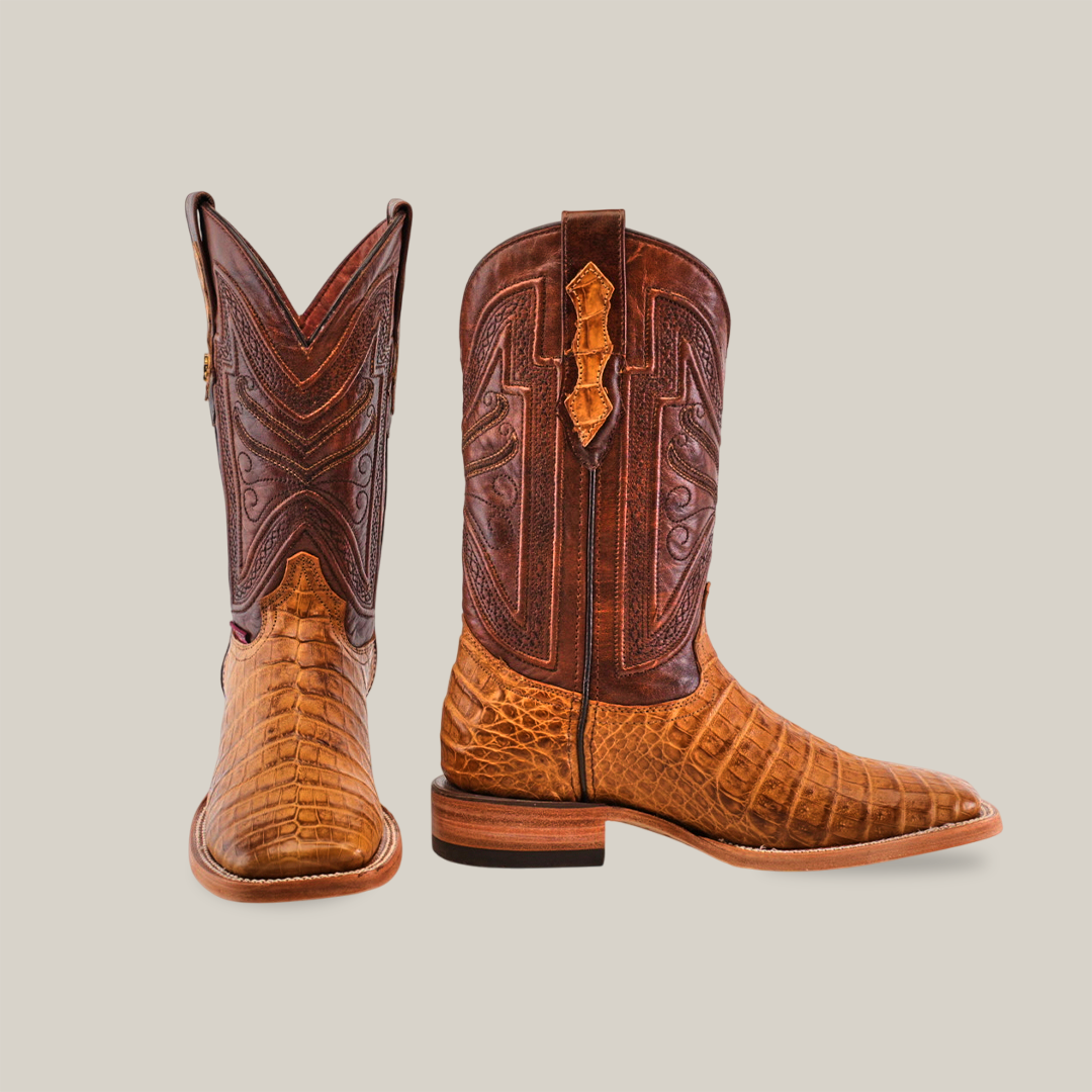 The Exotic Caiman Belly - Camel - Square Toe boots are crafted from rich brown caiman leather, featuring a textured crocodile skin on the foot and a smooth, patterned shaft. One boot stands upright, while the other angles to the right.