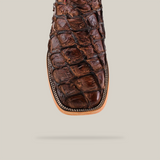 Close-up of the Exotic Pirarucu Fish brown cowboy boot featuring a square toe. The scaly texture complements Western style, outlined with white stitching against a light beige background.