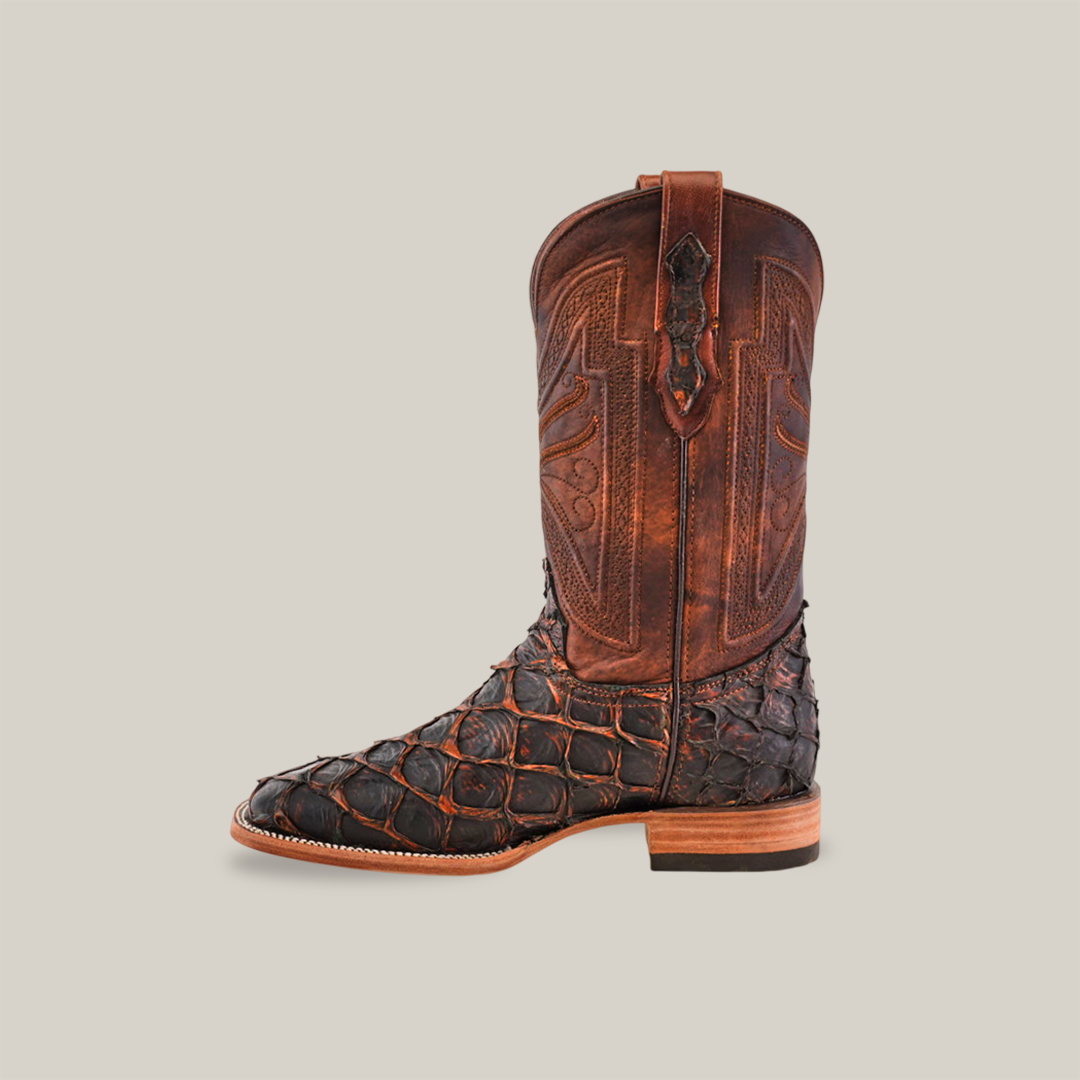 The Exotic Piraruco Fish boot in Rustic Cogñac features an intricate brown leather shaft and a textured dark-toned foot with a square toe. It has a medium heel, pull tab, and is elegantly displayed on a light beige background.