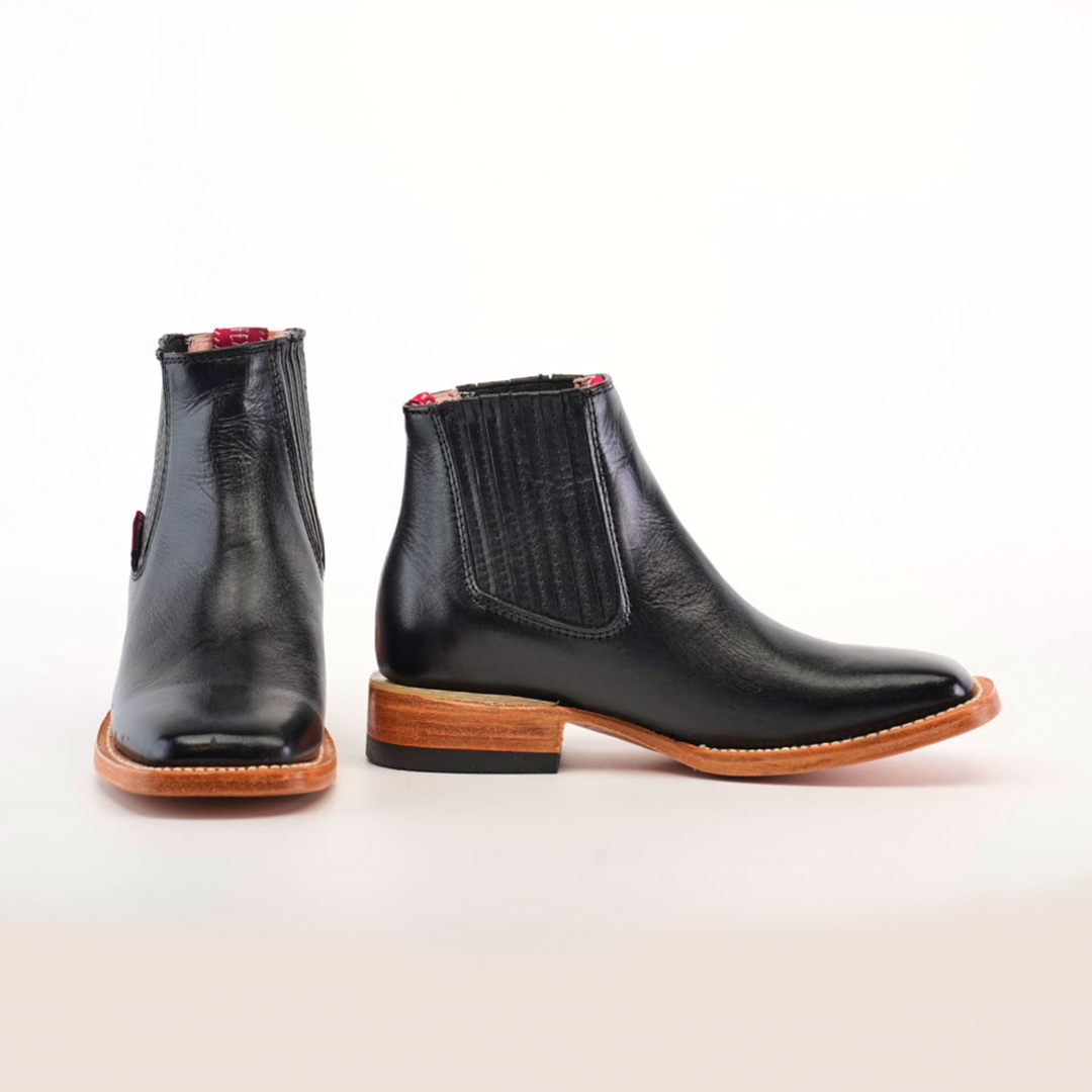 The Napa Black Square Toe boots feature wooden soles, with one boot showcasing the front and sole and the other at an angle to highlight its elastic side panel. The inside has a chic red pattern, making it ideal for any fashion-forward wardrobe.