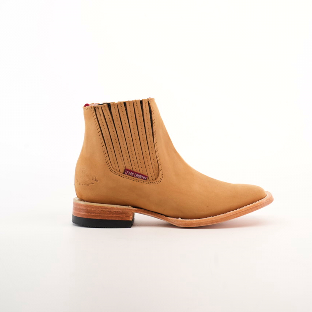 The Prime Suede Honey Square Toe boot showcases a tan leather body with a square toe, brown elastic side panels, and a black heel. Its lighter natural leather sole offers a stylish contrast against the upper, bringing a hint of cowboy flair. Its presented on a plain white background.