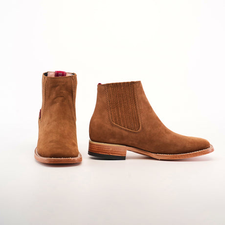 Two Prime Suede Caramel Square Toe ankle boots are displayed, one facing forward and the other showing the side, on a plain white background. They feature brown suede with classic design, wooden sole, elastic side panels, and a subtle square toe charm.