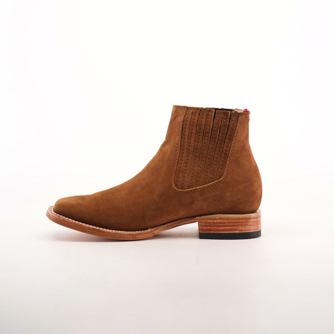 The Prime Suede Caramel Square Toe boots offer rustic charm with their brown suede finish, side elastic panel, decorative stitching, and square toe design. They feature a wooden sole paired with a black heel and are showcased on a plain white background.