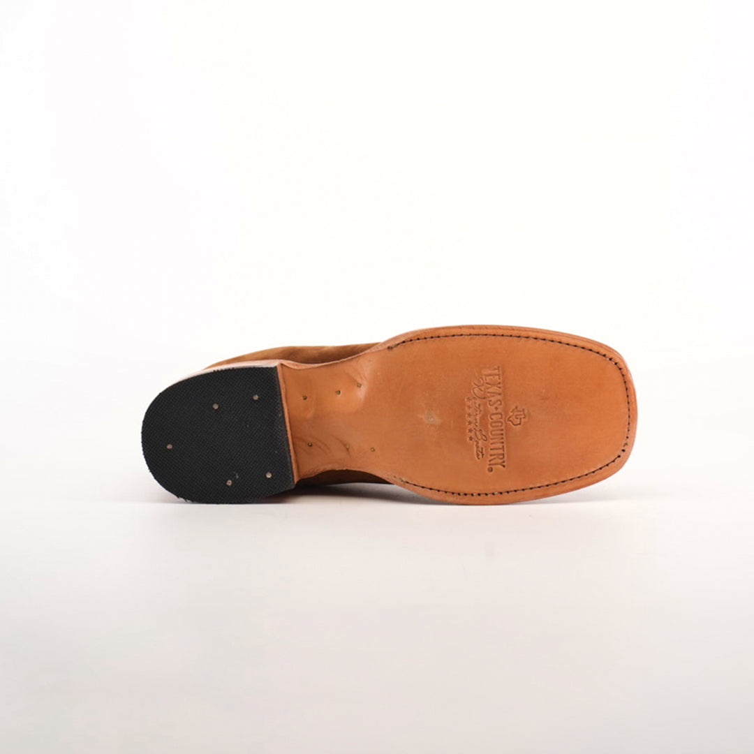 The image showcases the sole of a Prime Suede Caramel Square Toe shoe. It has a tan hue with To Boot New York branding, a square toe design, and a heel reinforced with black rubber and metal studs. The shoe is set against a plain white background.