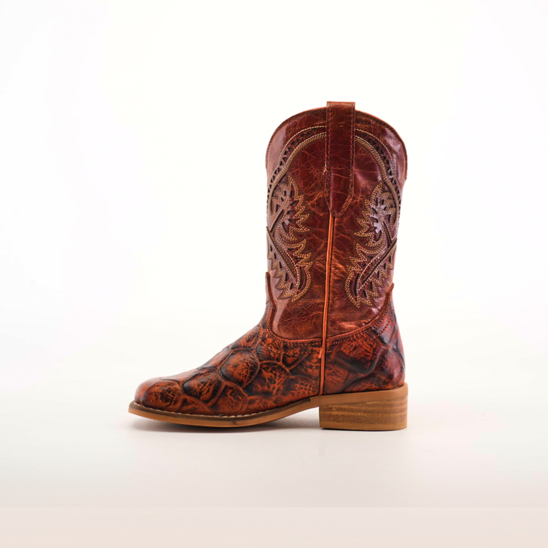 The Pirarucu Bass Print Cogñac Square Toe boot is a single brown cowboy boot showcasing ornate stitching and a textured Pirarucu bass print on the lower half. It features premium leather, a wooden heel, square toe, and pull tab, all set against a plain white background.