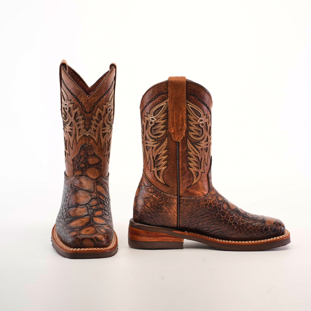 The Loggerhead Turtle Print brown Rodeo Toe boots are crafted from premium cowhide leather, featuring intricate stitching and a scaly texture, reminiscent of loggerhead turtle print. These childrens cowboy boots have a flat heel and are showcased against a plain white background.