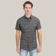 Against a plain background, a person models the Mens Performance Western Short Sleeve Aztec Print Black/Brown Shirt - PGW10635 paired with light blue jeans. With short dark hair and a neutral expression, the individual effortlessly exudes understated style with this geometric-patterned ensemble.