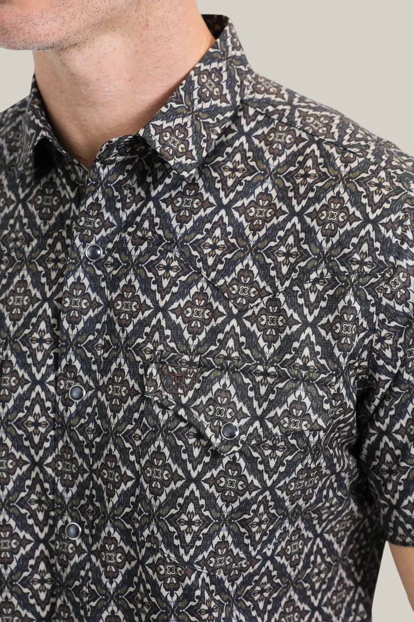 A person wears a Mens Performance Western Short Sleeve Aztec Print Black/Brown Shirt (PGW10635), featuring a geometric pattern with small flower motifs in diamond shapes and two buttoned chest pockets, exuding classic Aztec appeal.