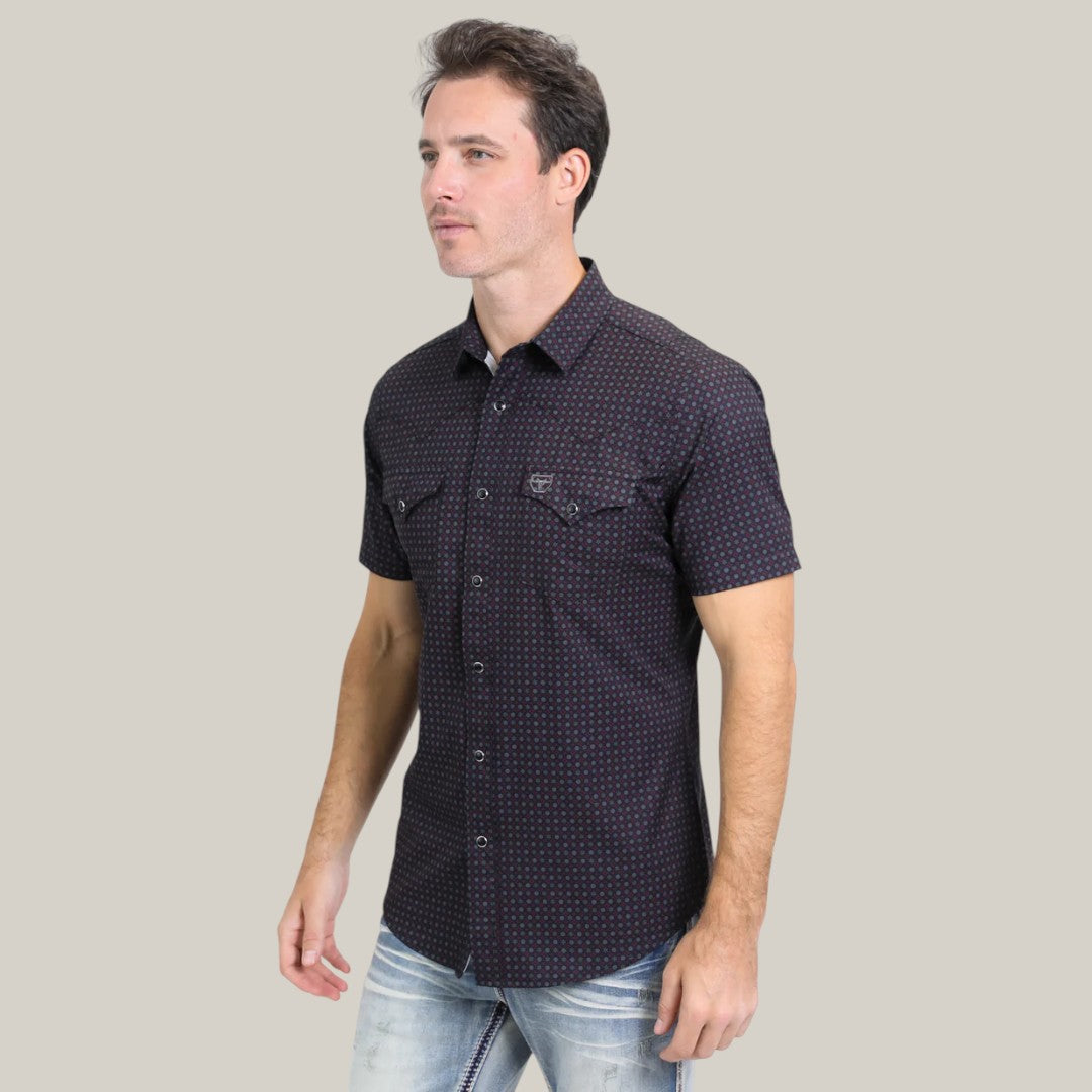 A person wearing the Mens Performance Western Short Sleeve Aztec Print Black Shirt (PGW10639) stands against a plain light background, gazing to the side with a neutral expression, paired with light blue jeans for a stylish, casual look.