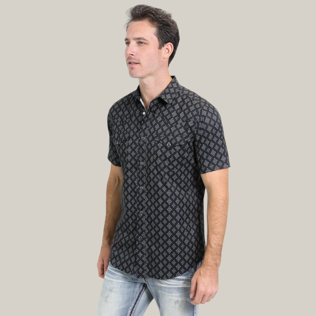 Standing against a plain backdrop, a person dons the Mens Performance Western Short Sleeve Aztec Print Black Shirt (PGW10630), crafted with quick-dry fabric for versatile occasions, paired with light blue jeans. With short, dark hair, they exude confidence while gazing slightly to the side.