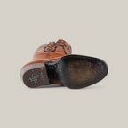 A California Tall Shaft Honey cowboy boot lays on its side, showcasing intricate sole designs. Its polished brown leather surface features branding and ornate patterns, while the round toe pairs beautifully with a plain, light gray background.