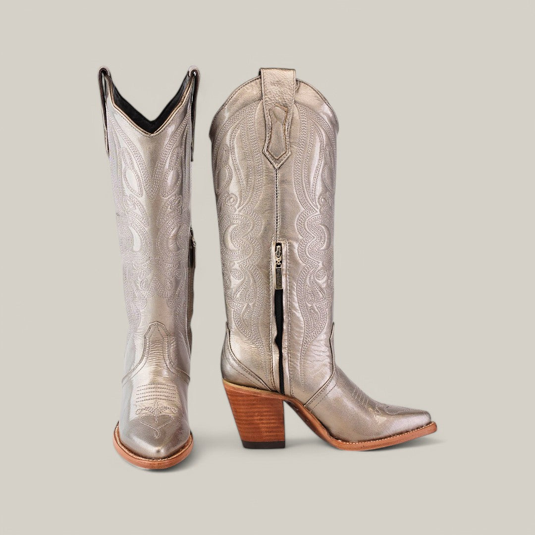 The Linda Tall Shaft Mercury - J Toe boots from the Platinum Collection are silver cowboy boots with intricate patterns and wooden heels. Featuring a side zipper, one boot stands upright while the other tilts slightly against a plain gray background, perfect for any stylish ensemble.