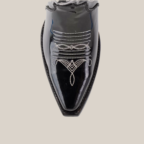 A close-up of the Artemisa Patent Black shoe reveals premium leather and intricate white stitching on the snip toe, forming elegant symmetrical patterns that highlight its craftsmanship against a plain light background.