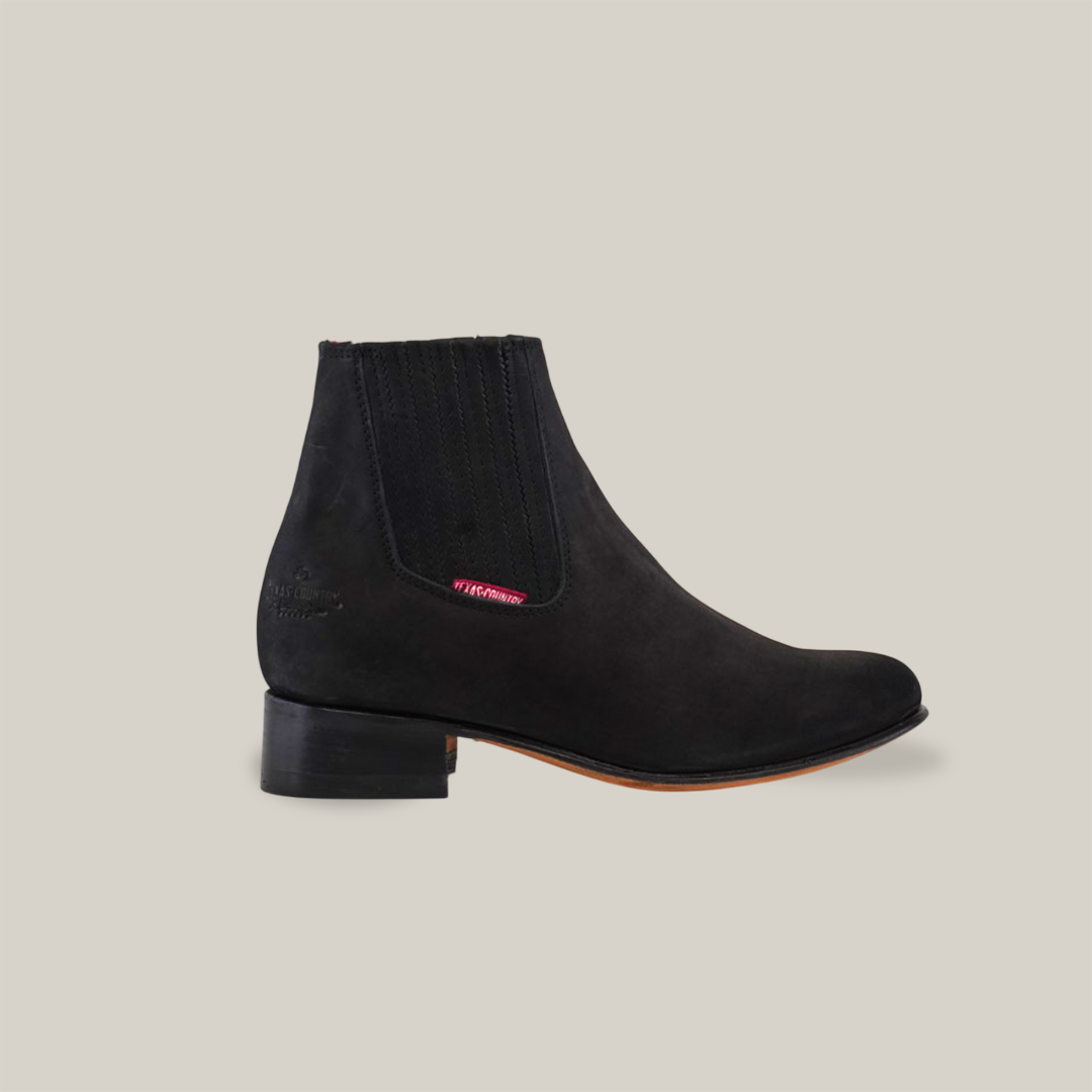 The Prime Suede Black Round Toe is a black suede ankle boot with round toe design and elastic side panels, featuring a distinctive reddish logo tag contrasting against the dark, glossy heel. Ideal for pairing with cowboy-inspired ensembles, shown on a plain white backdrop.