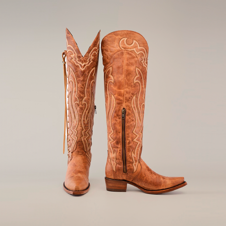 A pair of Tania Fawn cowboy boots with a tall shaft and snip toes features intricate embroidery, side zippers, and luxurious leather soles. One boot stands upright while the other is tilted, revealing wooden heels on a plain white background.