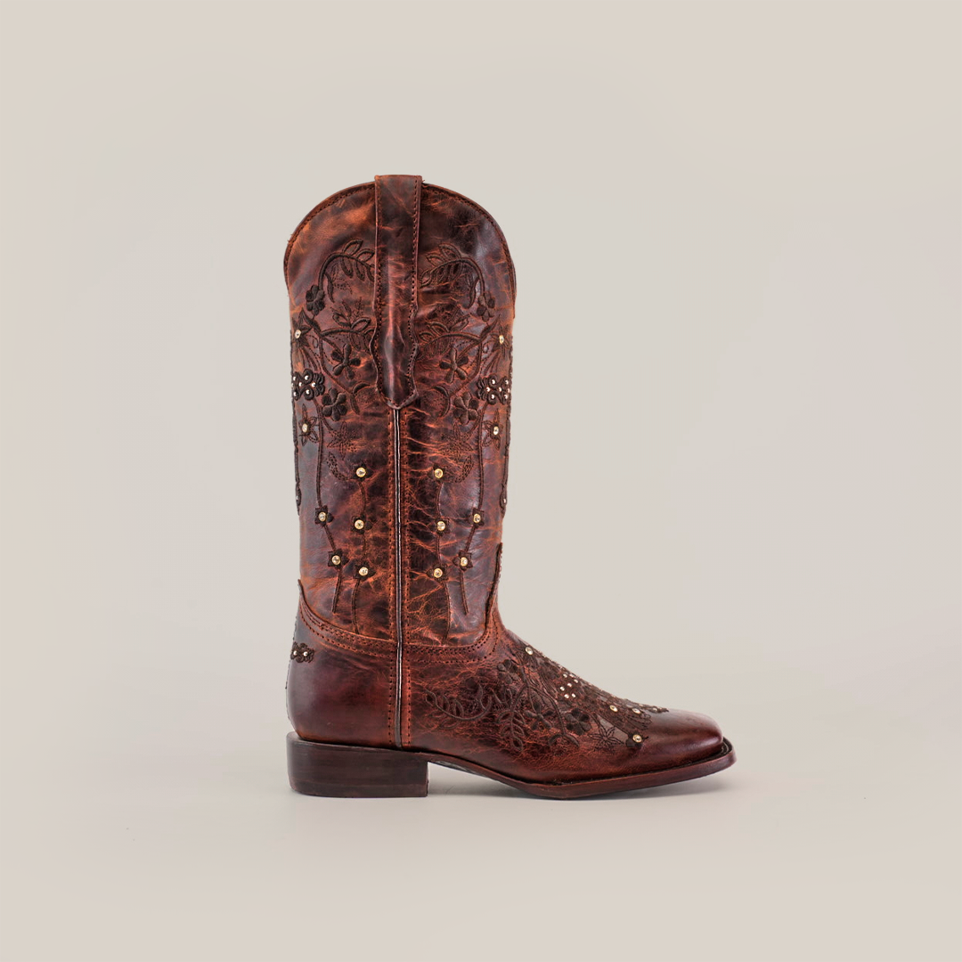 A single Marga Cruz Brick Mid Shaft boot, featuring intricate floral embroidery and gold studs, stands against a white background. Crafted from premium leather, its weathered look highlights the artistry of handcrafted boots.