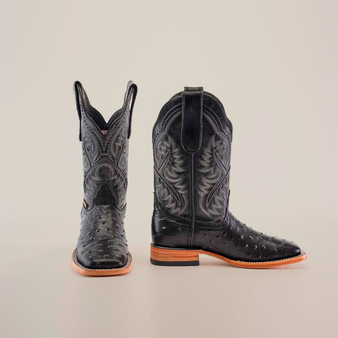 The Karoo Ostrich Print Black Square Toe boots feature a luxurious design with intricate patterns, a smooth, shiny texture, and exotic ostrich print. Displayed against a white backdrop, one boot stands upright while the other lies on its side, showcasing decorative stitching and a light brown sole.