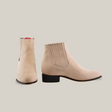 The Prime Suede Pearl Round Toe boots are shown in beige with a smooth finish. One boot faces forward while the other is turned sideways, highlighting the handcrafted stitched detail. They feature a low black heel and rounded toe.