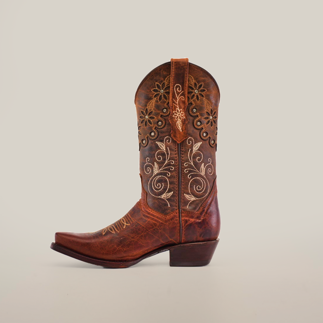 The Grecas Moka Crystals cowboy boot features intricate white floral embroidery and scroll patterns on a brown surface. This handcrafted luxury item showcases artisanal craftsmanship, highlighted by its snip toe and slight heel, all presented against a plain background.